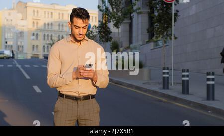 Stylish bearded hispanic businessman stand in city center street uses mobile phone texting scrolling tapping app technology online communication with Stock Photo