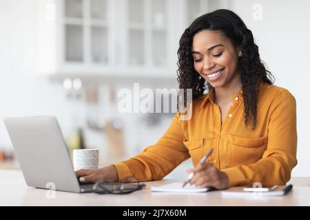 Cheerful pretty black lady working from home, taking notes Stock Photo