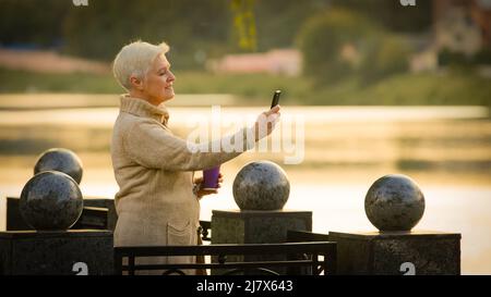 Contented middle-aged lady making video call looking at smartphone screen talking on cell phone. Elderly woman in retirement age taking photo on Stock Photo