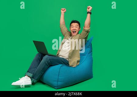 Overjoyed asian man making YES gesture, sitting in beanbag chair with laptop, celebrating success over green background Stock Photo