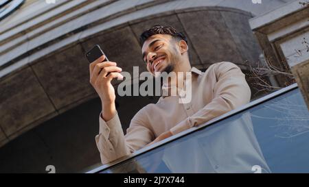Bottom view successful happy smiling arabic hispanic bearded millennial business male user client standing on balcony terrace at home office holding Stock Photo