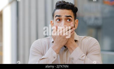 Confused afraid scared young guilty man shocked surprised guy male hispanic arab indian covers mouth with hands worried about telling secret feels Stock Photo