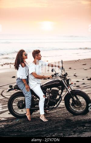 male and female couple in white outfit sitting on a motorcycle at a black sand beach in Bali Indonesia on a black vintage motorcycle during sunset Stock Photo
