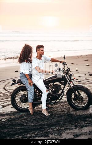 male and female couple in white outfit sitting on a motorcycle at a black sand beach in Bali Indonesia on a black vintage motorcycle during sunset Stock Photo