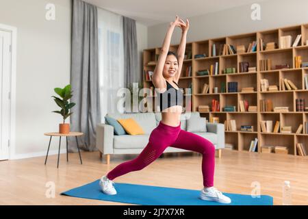 Young asian lady exercising at home, standing in warrior pose on fitness mat, copy space, full length shot Stock Photo