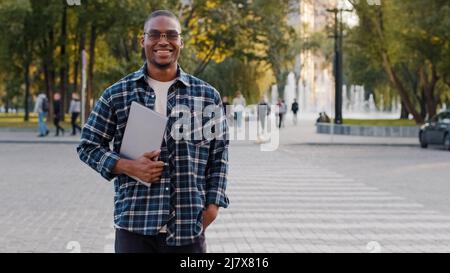 Young successful african american guy wearing sunglasses male business man with laptop smiling stands against crosswalk road traffic background Stock Photo
