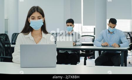 Multiethnic staff sit at coworking space with laptops during workday in modern office. Pensive girl busy with work task. Wear facial masks, keeping Stock Photo