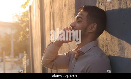 Side view handsome bearded hispanic arabic business man client guy standing near stone wall talking on mobile phone answering call talks with Stock Photo