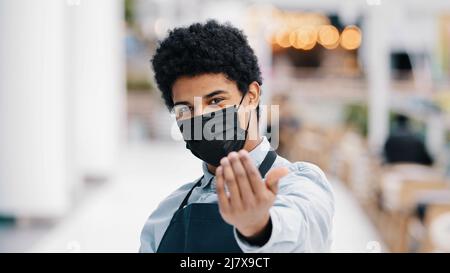 Friendly african masked guy male waiter salesman man in medical face protective mask restaurant cafe kitchen bar worker pointing hey you at camera Stock Photo