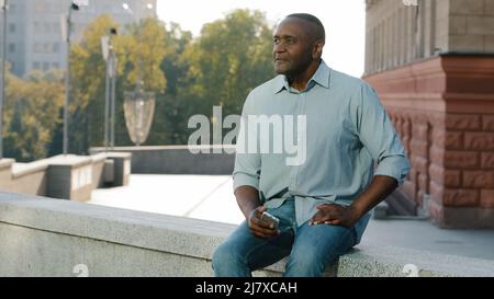 Stressed frustrated elderly African American man of retirement age in casual clothes shirt and jeans sitting outdoor holding phone waiting for client Stock Photo