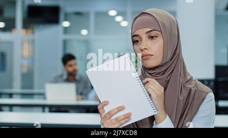 Thoughtful millennial indian woman in hijab looking aside, planning preparing for meeting. Pensive young Arabic female worker sitting at office using Stock Photo