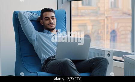 Relaxed happy Indian businessman student worker sits in comfortable armchair meditating, positive calm Arabic man relax break hold hand behind head Stock Photo