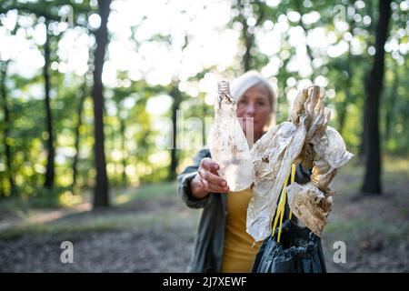 Senior woman ecologist with bin bag picking up waste outdoors in forest. Stock Photo