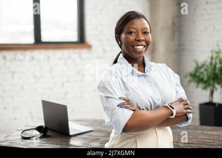 High-skilled African-American businesswoman wearing smart casual shirt stands with arms crossed in modern office space. Multiracial bossy lady, purposeful leader, female entrepreneur Stock Photo