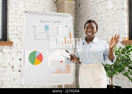 Modern African-American female business coach, college tutor or trainer looking at webcam and standing near whiteboard, talking to online audience, giving virtual online lesson, educational course Stock Photo