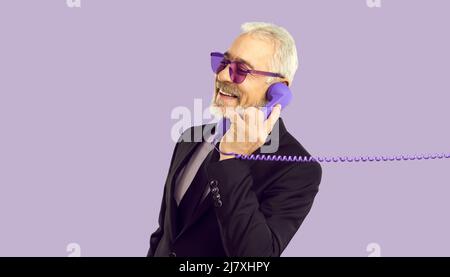 Cheerful stylish and charismatic senior man talking on landline phone on purple background. Stock Photo