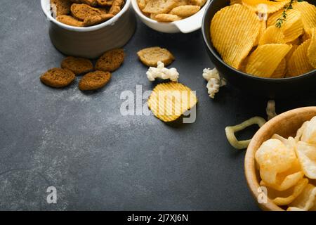 Unhealthy food. Snacks. All classic potato snacks with peanuts, popcorn and onion rings and salted pretzels in bowl plates on black concrete backgroun Stock Photo