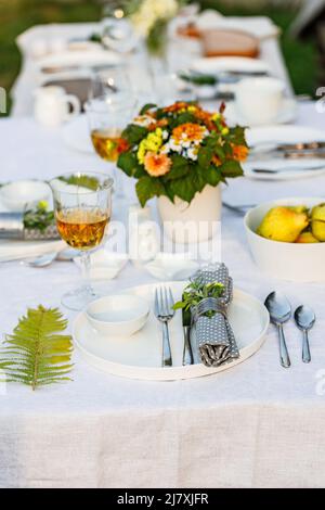 Elegant table setting with white dishes on a white linen tablecloth. Fruits, drinks in crystal glasses. Summer holiday table set up. Outdoor eating on Stock Photo