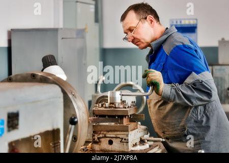 Elderly turner of 50-55 years old in overalls works at lathe in production shop. Processing of metal products. Real portrait of Caucasian worker. Production background. Stock Photo