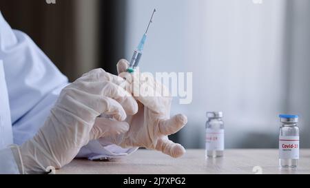 Unrecognizable doctor nurse medical worker in white coat and latex gloves holds syringe splash medicine preparing for injection sitting at table with Stock Photo