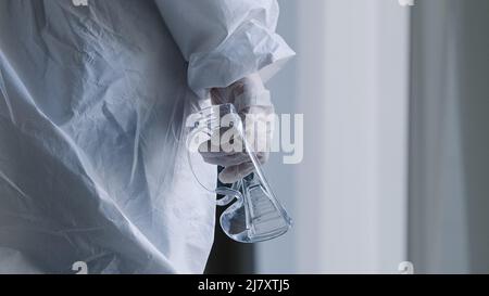 Close-up unrecognizable doctor unknown nurse surgeon practitioner in white uniform protective suit in latex medical gloves holds transparent glasses Stock Photo