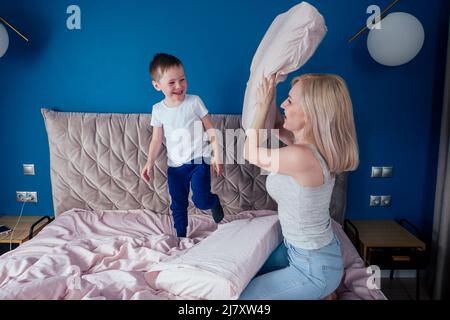 Family having fun together at the weekend together.Mother and child on bed. blonde mom and baby boy playing pillow fight in morning bedroom. Parent Stock Photo