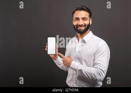 Mock-up concept. Handsome confident bearded Indian man in formal wear showing smartphone with empty screen, points finger at blank device display isolated on black, male advertising new mobile app Stock Photo