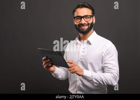 Modern Indian businessman in formal wear using digital tablet isolated on black. Portrait of male office employee using online technology for doing business, computer app for accounting Stock Photo