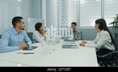 Young indian team leader sitting with colleagues, discussing issues at negotiations meeting. Skilled millennial diverse employees working together at Stock Photo