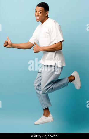 Excited overjoyed African-American young guy jump up on blue background, having fun in studio, cheerful man wearing casual t-shirt and jeans celebrating good news, victory, jumping for happiness Stock Photo