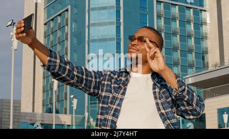 Stylish happy african man in trendy sunglasses speaks webcam mobile phone in city. American male blogger vlogger guy video call making photo using Stock Photo