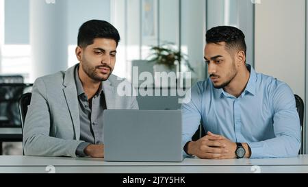 Two serious focused Arab young male businessmen sitting in modern office using laptop looking at computer screen. Diverse employees East Indian Stock Photo