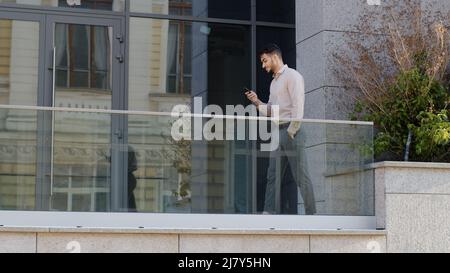 Successful business man boss leader worker manager well-dressed guy walks on balcony terrace in company office looking at mobile phone chatting online Stock Photo
