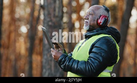 Elderly african american forestry engineer in noiseisolating headphones in forest old mature foreman forester thinks over work plan for cutting trees Stock Photo