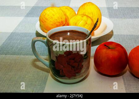 A mug of cappuccino is on the table. In the background, oranges without zest on a white plate. Red apples next to the cup. Stock Photo