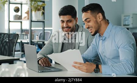 Cheerful positive friendly businessmen partners colleagues holding document contract looking at laptop screen discuss ideas having fun during break in Stock Photo