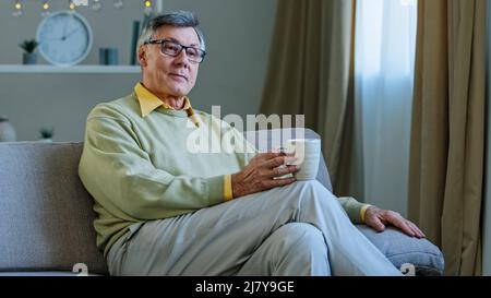 Old man wearing glasses sit on couch in living room hold hot drink cup pensive elderly grandpa relaxing alone at home drinking tea looking into Stock Photo