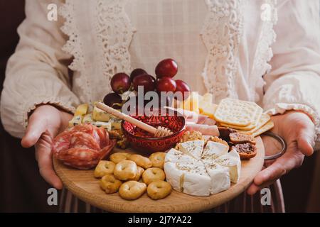 A woman holds in her hands a round wooden cutting board with Wine appetizers - slicing cheese, sausage and fruit. Stock Photo