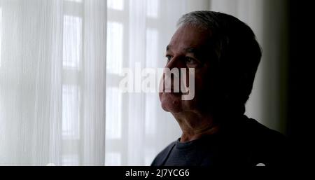 Contemplative senior man by home window looking outside Stock Photo