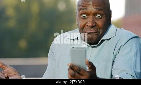 Mad stressed african man holding cellphone annoyed with mobile app error spam message on slow stuck broken smartphone, angry black person frustrated Stock Photo