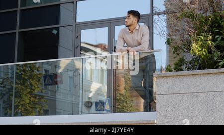 Confident young hispanic business man standing at railing modern skyscraper office building looking away holding phone thinking resting. Successful Stock Photo