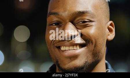 Male happy portrait african american young millennial satisfied man stands in city in evening town lights background looking at camera smiling toothy Stock Photo