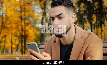 Portrait young serious guy use app hispanic man looking at phone screen browses news on telephone concentrated male sits on bench in autumn park read Stock Photo
