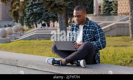 American student guy african business man sitting outdoors in city street typing on laptop working online with computer e-learning listening to music Stock Photo