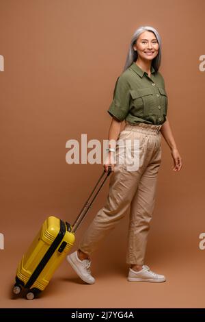 Full length photo of smiling senior caucasian woman in casual clothes standing with yellow suitcase isolated on brown background. Travel and vacation concept Stock Photo