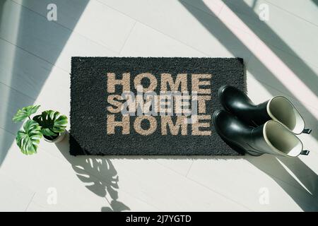 Rain boots on home sweet Home door mat during fall rainy season. Entrance doormat of house homeowner. Top view of rug for floor protection of rain Stock Photo