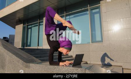 Strong acrobat business woman funny muslim girl in hijab stands on hands in acrobatic yoga pose asana balance handstand in city outdoors typing laptop Stock Photo