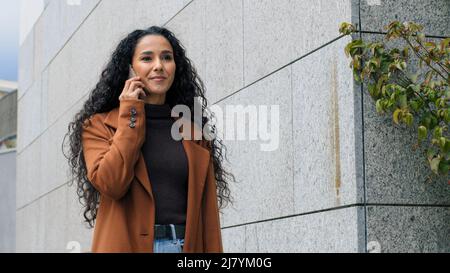 Portrait happy hispanic female latin girl business woman remote distant talk answering call walking in city talking on mobile phone on street. Lady Stock Photo