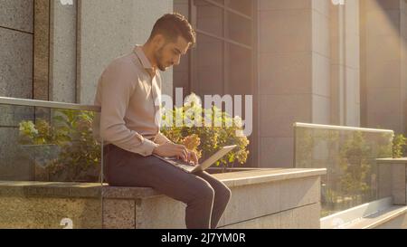 Hispanic businessman sitting outside with laptop working remote distant job outdoors near office company building. Young Arabian user student worker Stock Photo