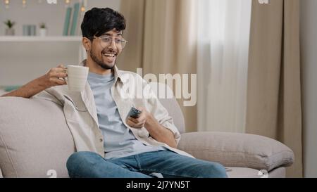 Arab man bearded guy with glasses resting at home sitting on couch drinking tea coffee watching funny movie championship sports game TV rejoices at Stock Photo